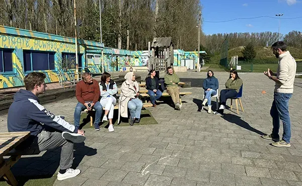 Foto van deelnemers aan de teambuilding luisterend naar de coach buiten op een bank in de zon